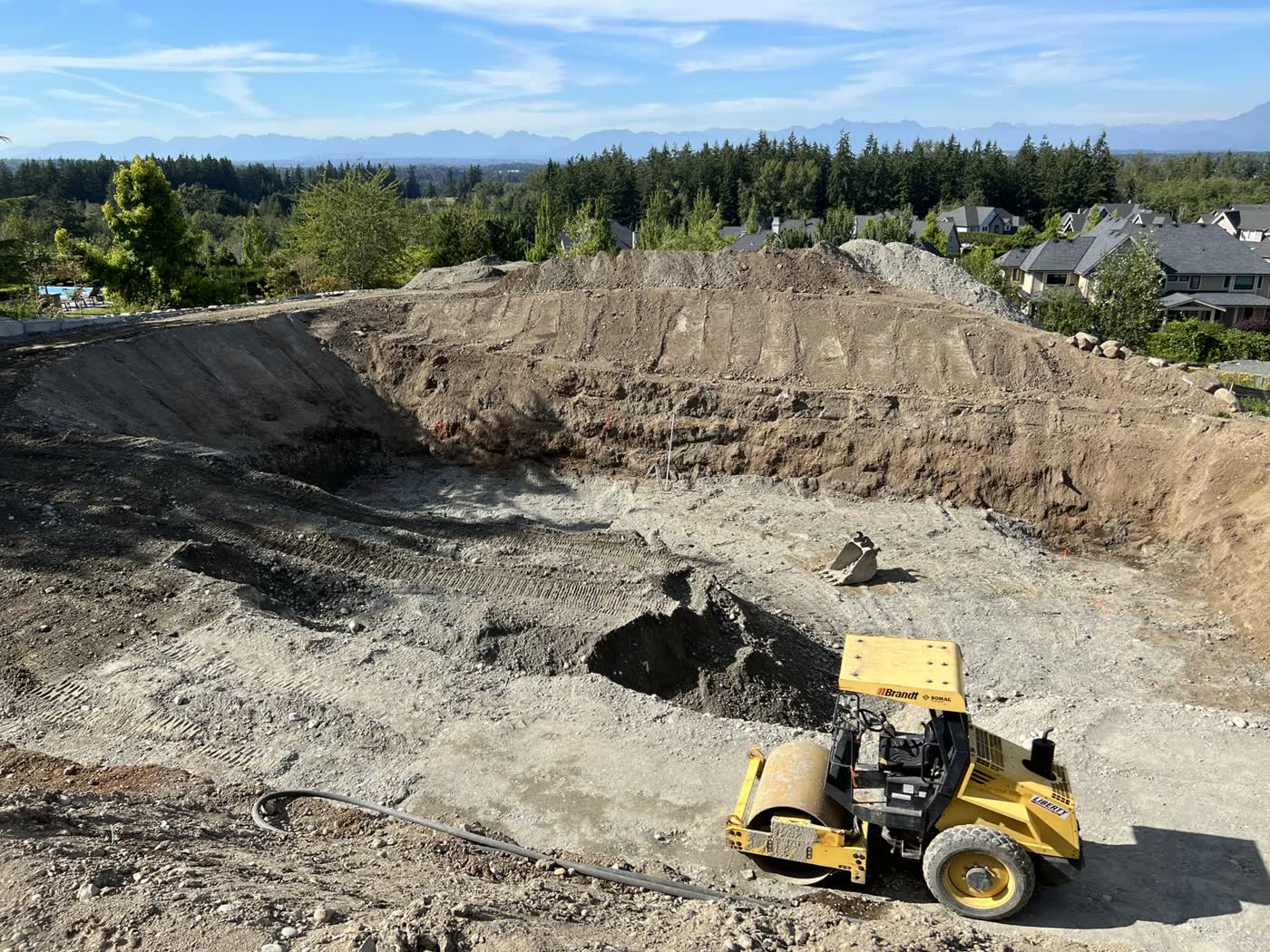A large excavation for a new home on a view lot, with a smooth drum roller compacting the floor of the dig and mountains on the horizon.