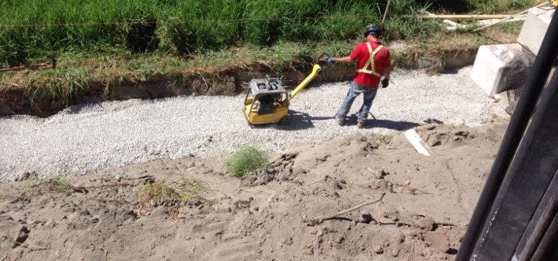 A crew member running a plate compactor over drain rock behind a new lock block retaining wall.